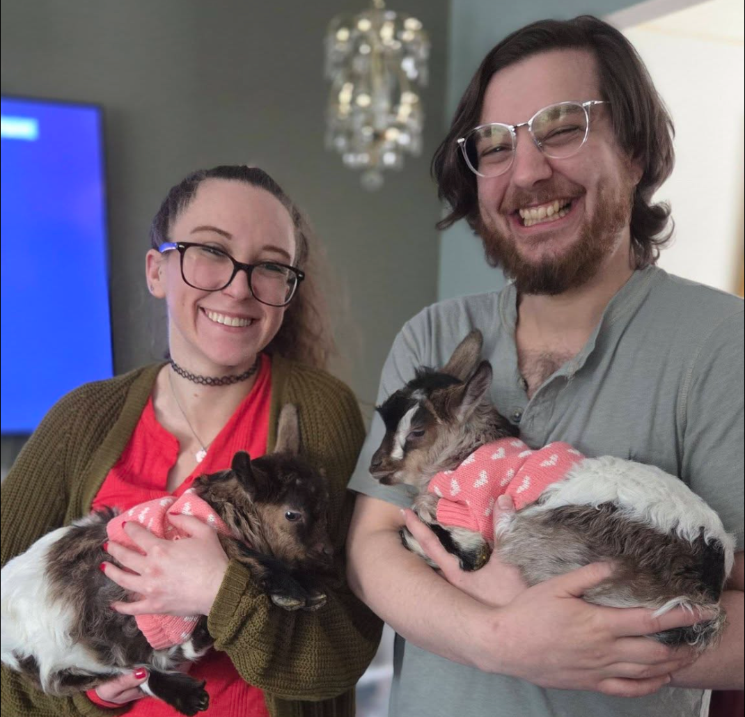 Two smiling visitors holding baby goats in pink sweaters