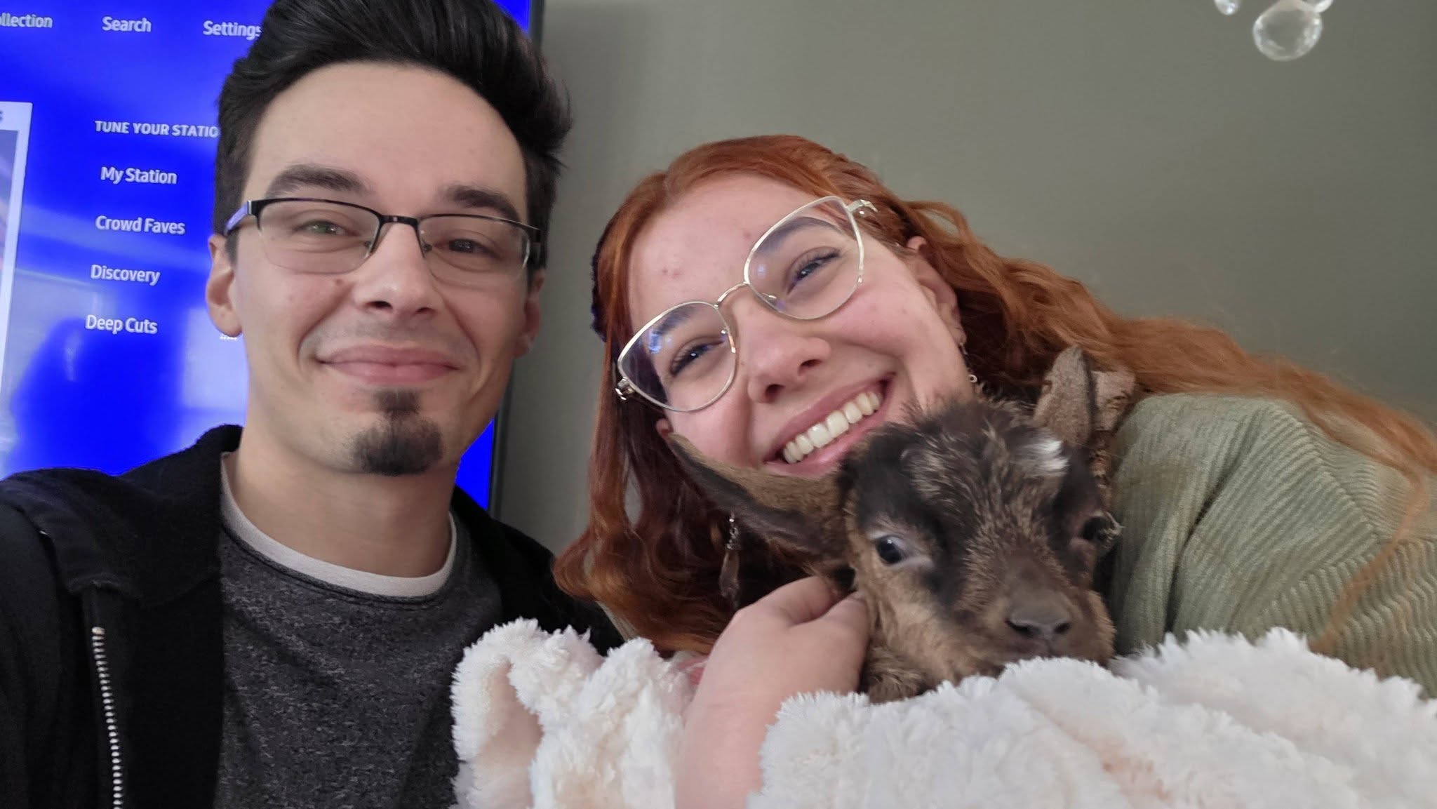 Two visitors smiling with a baby goat tucked into a blanket